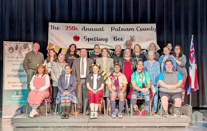 Three rows of people posed on a stage like a class photo. Behind them, a banner than reads "The 25th Annual Putnam County Spelling Bee"